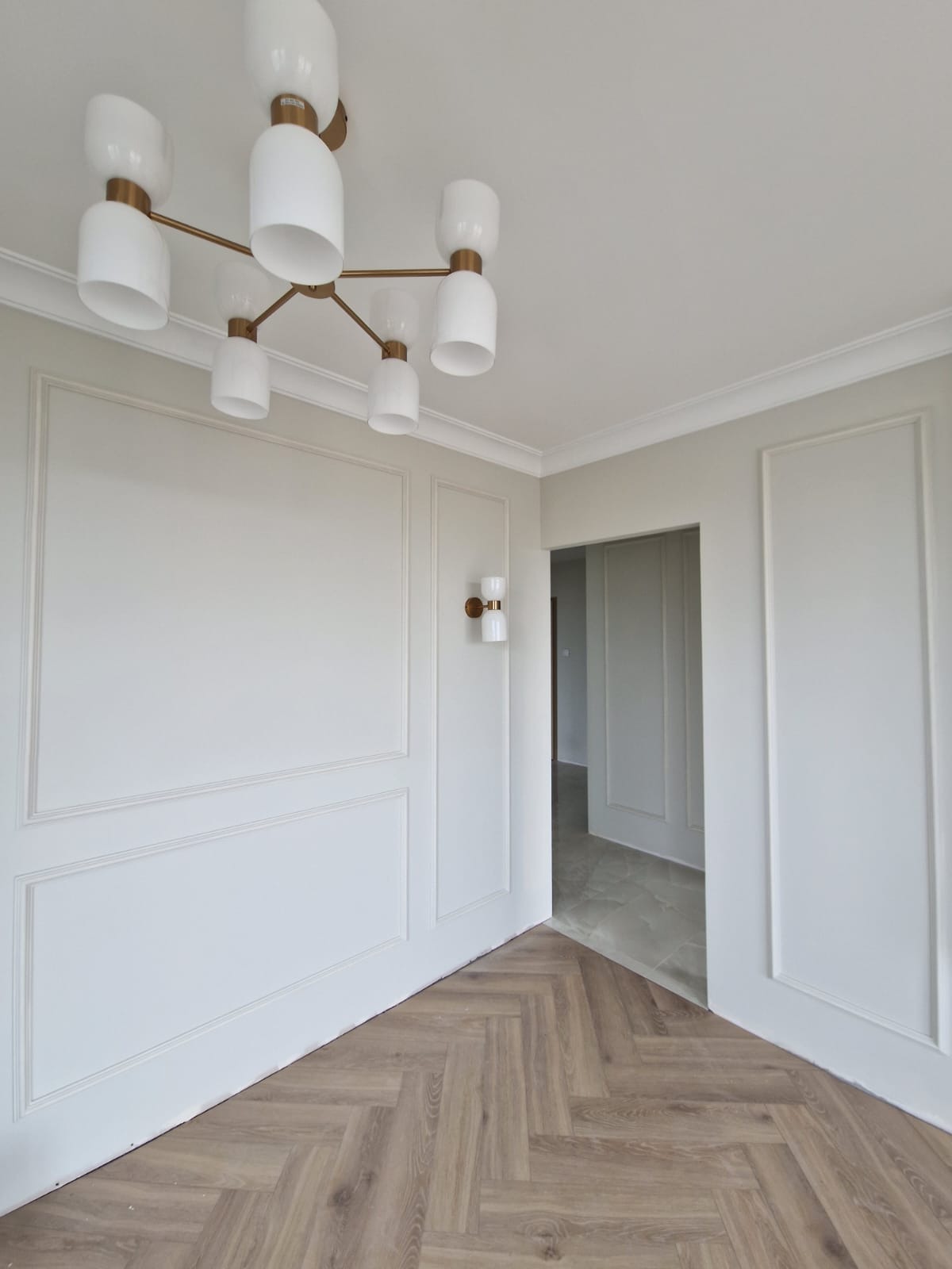 Jasne wnętrze po wykończeniu: parkiet w jodełkę, białe ściany z dekoracyjnymi listwami, nowoczesna lampa sufitowa i kinkiet. Minimalistyczny design.