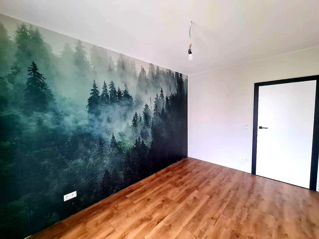 Pokój po remoncie: fototapeta z motywem lasu na ścianie, drewniana podłoga, białe ściany i drzwi z czarną ramą. Wisząca żarówka dodaje surowego charakteru.