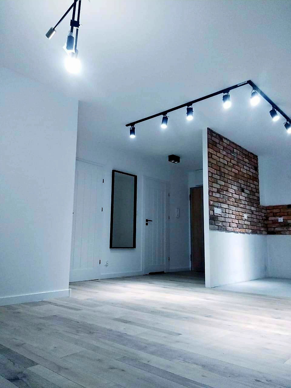 Wykończone wnętrze: jasna podłoga, białe ściany, ceglana dekoracja, oświetlenie szynowe. Minimalistyczny design z lustrem i drzwiami.
