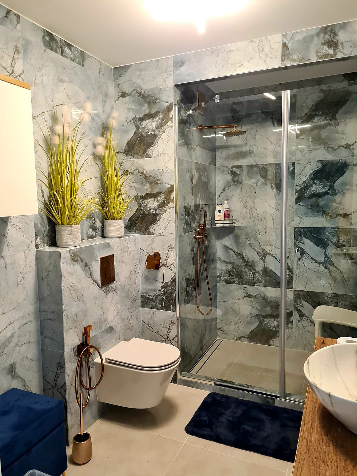 Stylowa łazienka z marmurowymi płytkami, kabiną prysznicową ze złotymi akcentami, wiszącą toaletą i roślinami w doniczkach. Nowoczesny design z dbałością o detale.