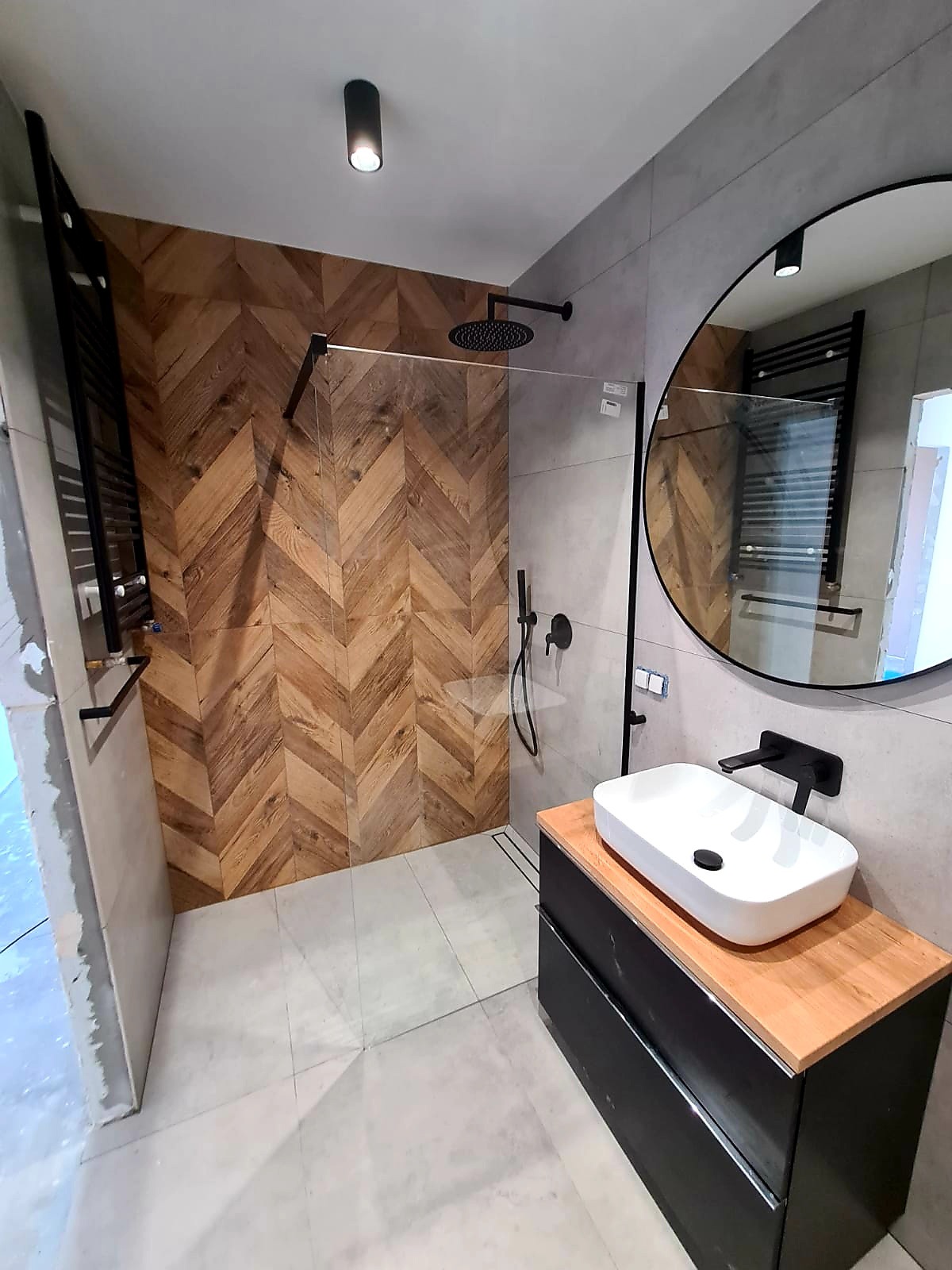 Nowoczesna łazienka z kabiną typu walk-in, drewnianą ścianą w jodełkę, okrągłym lustrem i białą umywalką na czarnej szafce z drewnianym blatem.
