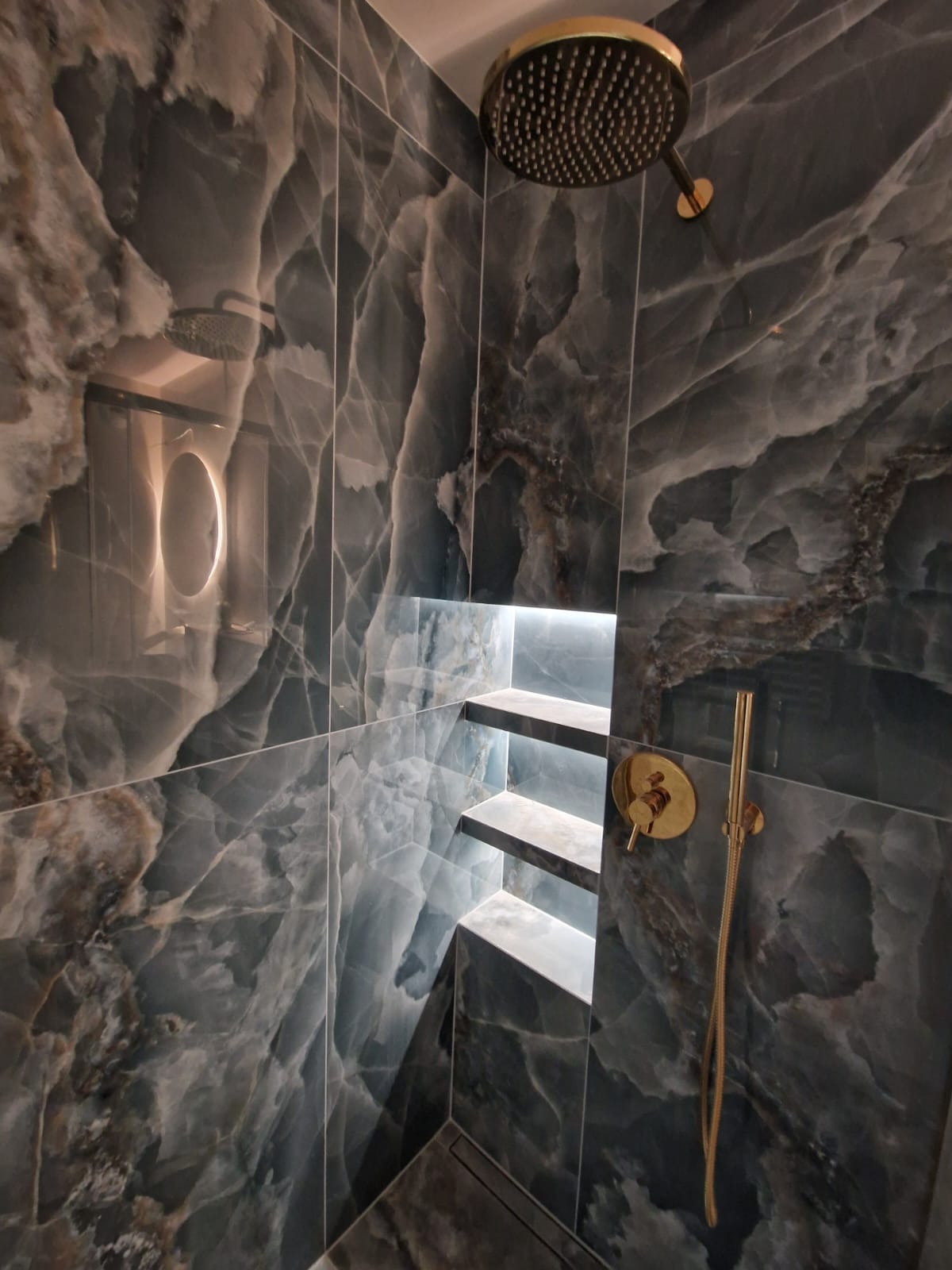 Luksusowa kabina prysznicowa z marmurowymi płytkami, złotą armaturą i podświetlanymi półkami wnękowymi. Nowoczesny design z dbałością o detale.