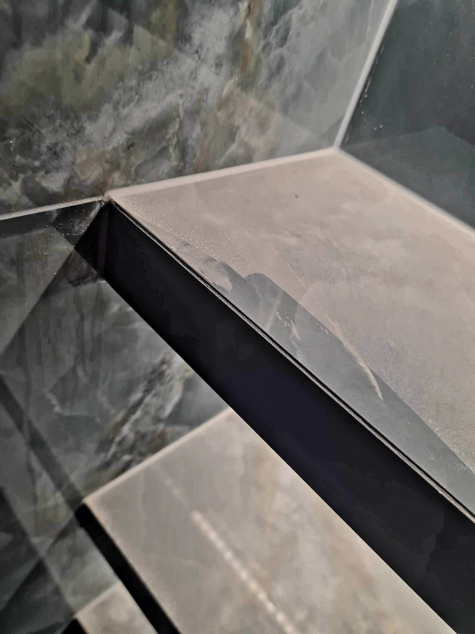 Półka z płytek imitujących marmur, wykończona czarną listwą, zamontowana w rogu. Widoczne detale fugi i wzoru płytek. Ujęcie z bliska, podkreślające precyzję wykonania.