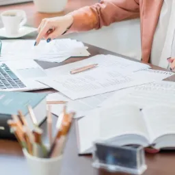 Kobieta w różowym żakiecie wskazuje palcem na dokumenty leżące na biurku obok laptopa, otwartej książki, filiżanki kawy i przyborów do pisania.