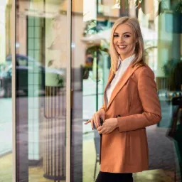 Portret uśmiechniętej blondynki w pomarańczowej marynarce, stojącej przed budynkiem z przeszkloną fasadą, w tle widoczny fragment ulicy i samochód.