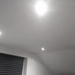 Biały sufit poddasza z czterema punktowymi lampami LED, widoczne okno z żaluzjami.