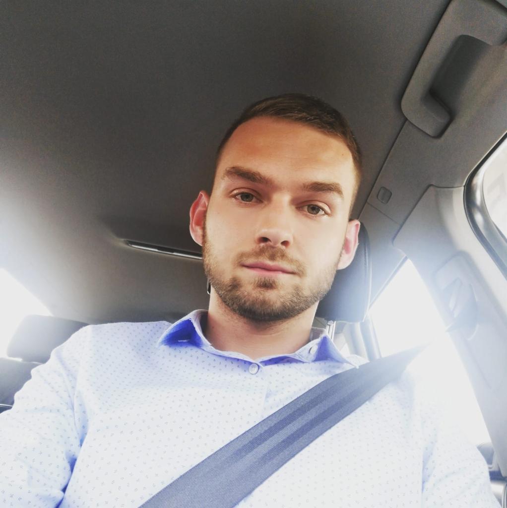 Młody mężczyzna w samochodzie, zapięty pasami, w koszuli z delikatnym wzorem. Widoczna tapicerka auta i fragment zagłówka.