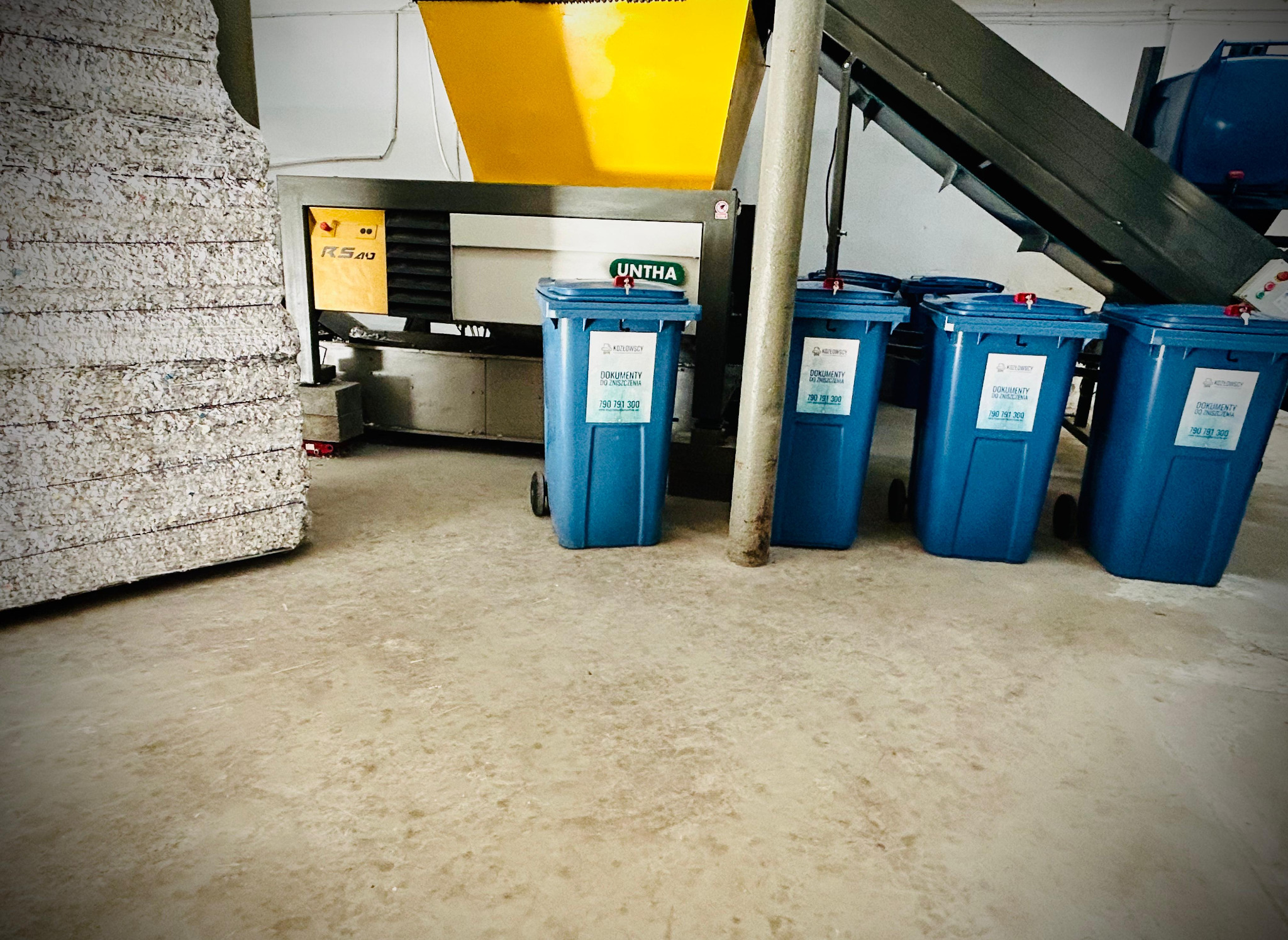 Stos pociętych dokumentów obok przemysłowej niszczarki UNTHA i rzędu niebieskich pojemników z naklejkami informującymi o usłudze utylizacji dokumentów firmy Kozłowscy.