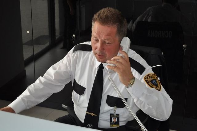 Mężczyzna w uniformie ochroniarza rozmawia przez telefon stacjonarny w pomieszczeniu z ciemnymi, szklanymi ścianami.