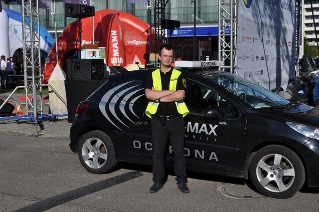 Mężczyzna w kamizelce odblaskowej stoi przed czarnym samochodem z napisem 'MAX SECURITY OCHRONA' na tle sceny z nagłośnieniem i dmuchanym łukiem.