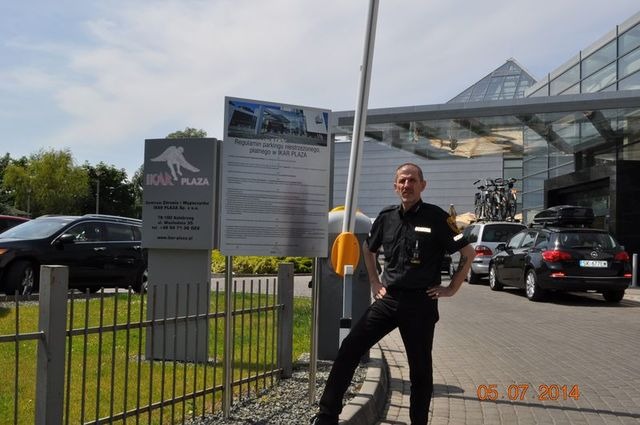 Ochroniarz w czarnym uniformie stoi na tle parkingu IKAR PLAZA z regulaminem parkingowym i szlabanem, w tle zaparkowane samochody.