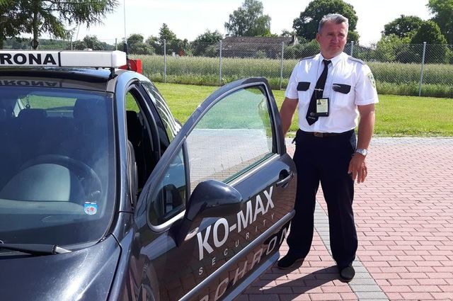 Mężczyzna w uniformie ochroniarza stoi obok otwartych drzwi samochodu z napisem 'KO-MAX SECURITY OCHRONA', na dachu pojazdu napis 'OCHRONA'.