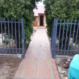 Nowo ułożony chodnik z kostki brukowej w odcieniach czerwieni i szarości, prowadzący do domu z czerwonym dachem, otoczony zielonymi tujami i metalowym ogrodzeniem w kolorze grafitowym, widok...