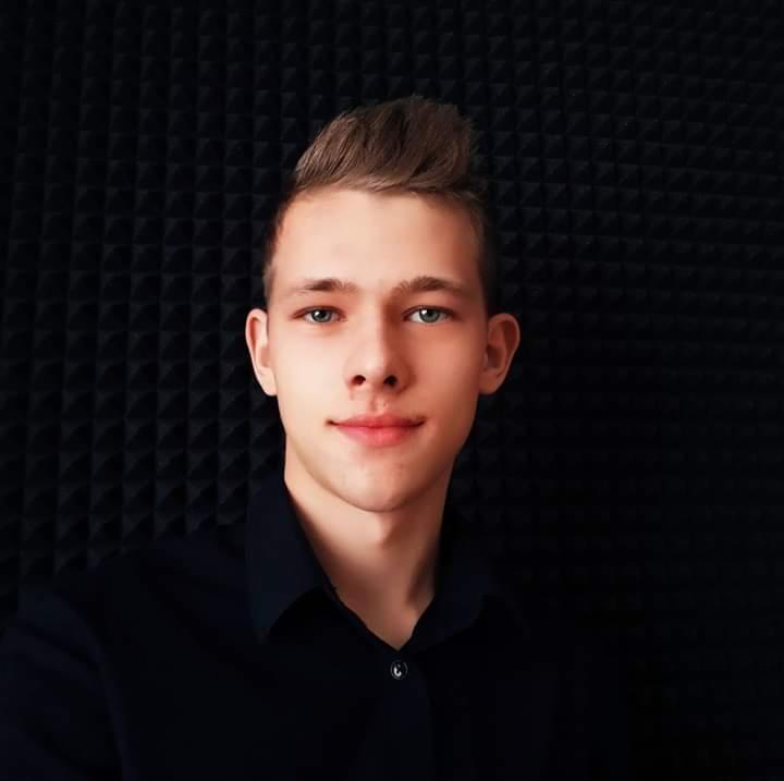 Portret młodego mężczyzny z krótkimi, zaczesanymi włosami, w ciemnej koszuli na tle czarnej, teksturowanej ściany.