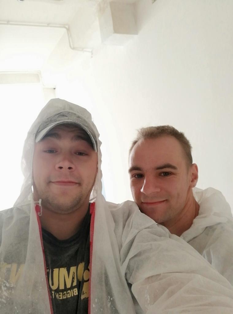 Dwóch mężczyzn w kombinezonach ochronnych, prawdopodobnie po pracy, uśmiechniętych na tle białej ściany.