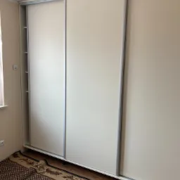 Trzydrzwiowa, biała szafa przesuwna z aluminiowymi prowadnicami, widoczna w pokoju na tle dywanu i ściany.