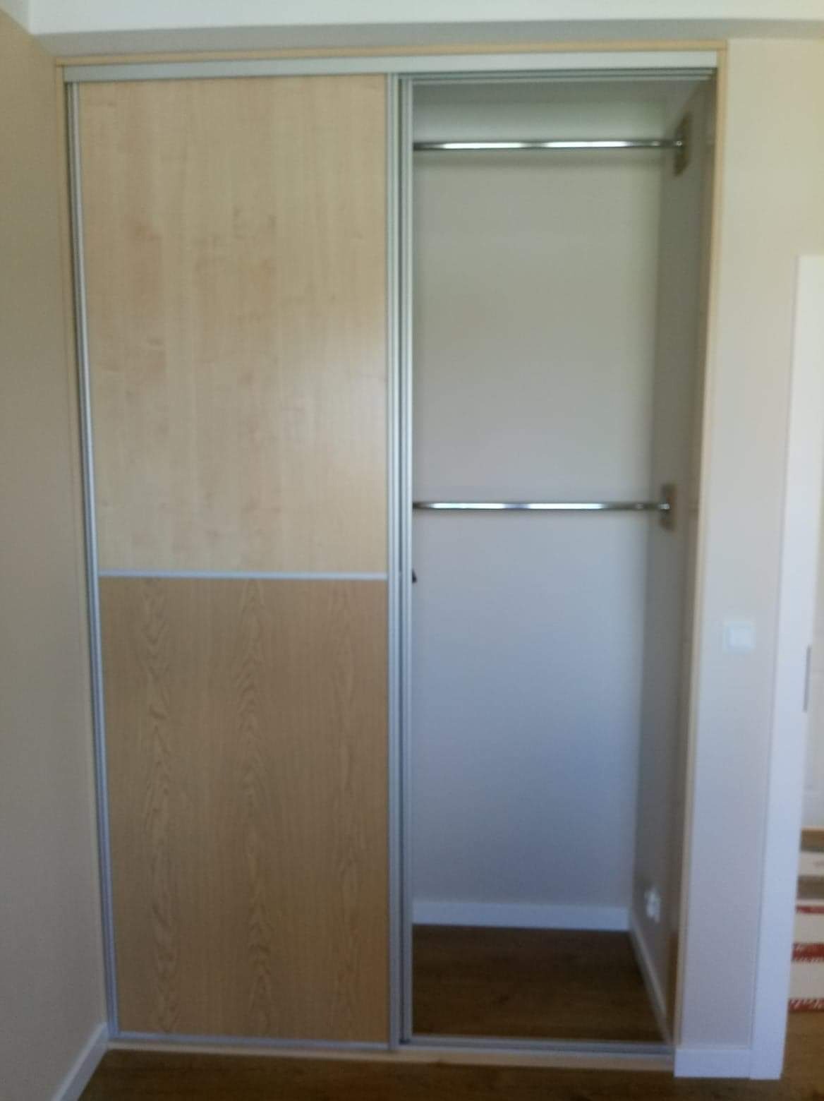 Wbudowana szafa z przesuwnymi drzwiami z jasnego drewna, jedna część drzwi zamknięta, druga odsłania wnętrze z dwoma drążkami na ubrania.