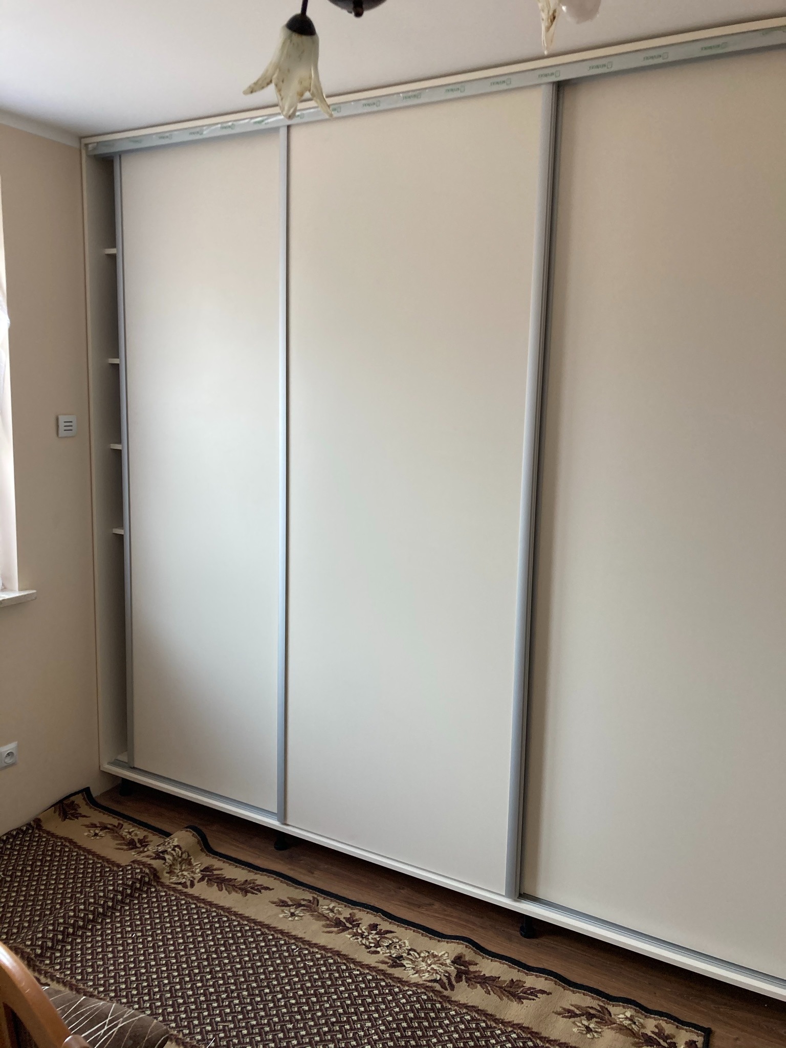 Trzydrzwiowa, biała szafa przesuwna z aluminiowymi prowadnicami, widoczna w pokoju na tle dywanu i ściany.