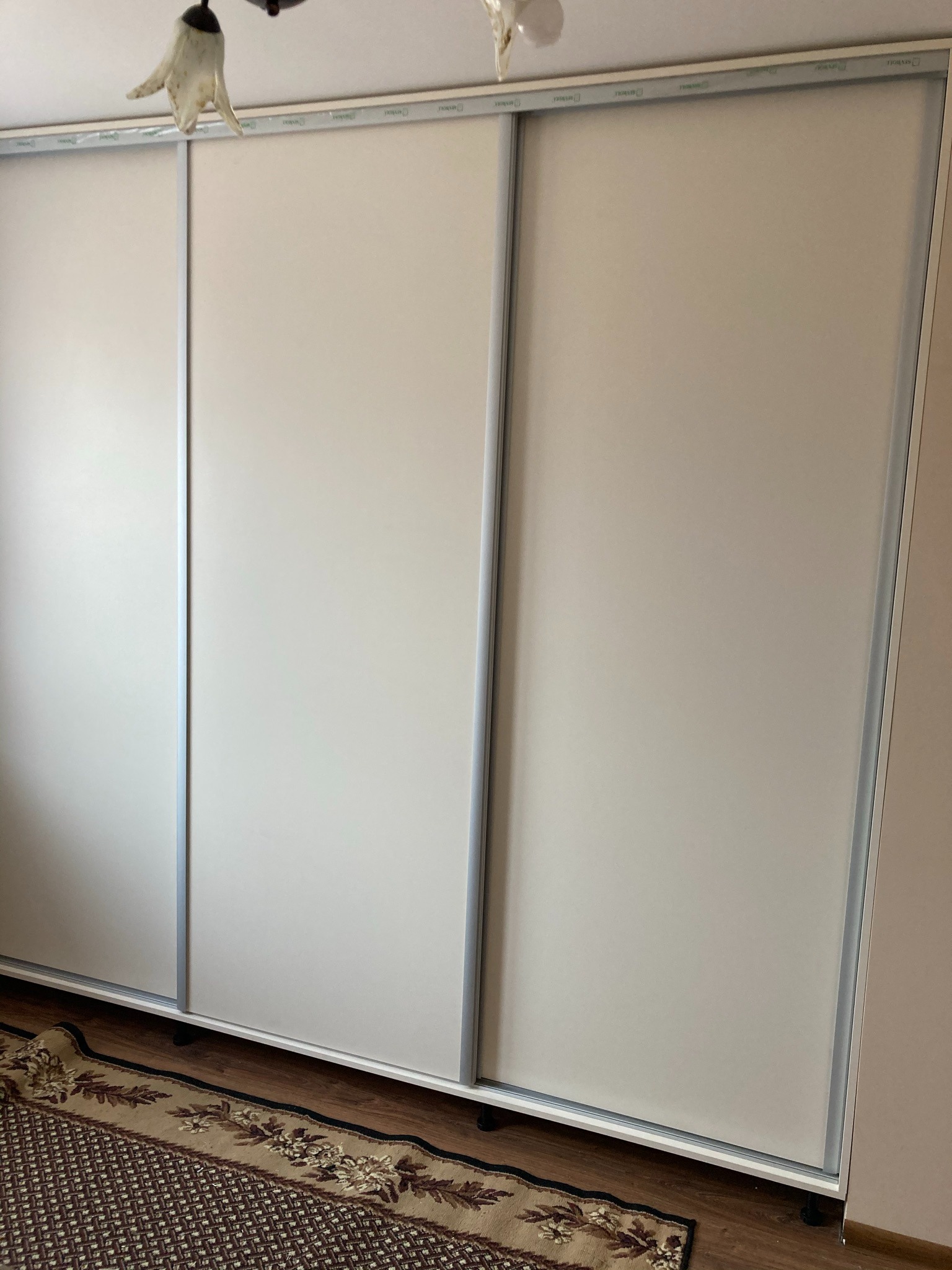 Trzydrzwiowa szafa przesuwna z białymi frontami i srebrnymi obramowaniami, widziana z perspektywy podłogi, z wzorzystym dywanem u dołu.