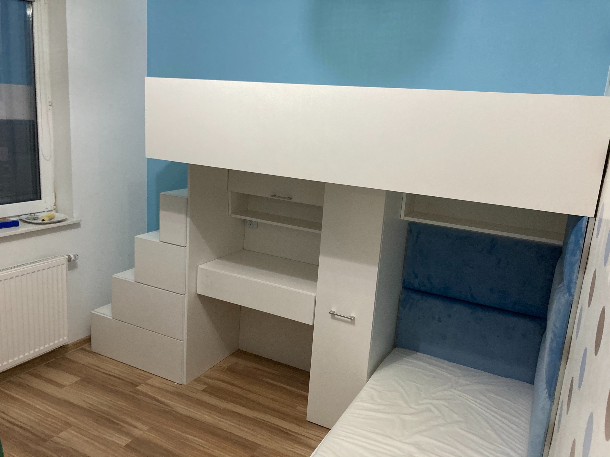 Białe łóżko piętrowe z biurkiem i schodami z szufladami, niebieska ściana w tle, widoczny fragment okna i grzejnika.