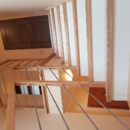 Nowoczesne schody z drewnianą ramą i metalowymi prętami balustrady, widok z dołu, jasne drewno, białe wypełnienia, ciemna szafka w tle.