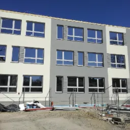 Trzypiętrowy budynek po częściowym remoncie elewacji, z szarą i białą kolorystyką, okna z odbiciem nieba, widoczne rusztowania i teren budowy z tablicą informacyjną 'Wyjazd z budowy'.