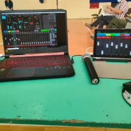 Laptop i tablet z oprogramowaniem do miksowania dźwięku, mikrofon na tle sali z publicznością, ustawienia korektora i kompresora widoczne na ekranach.