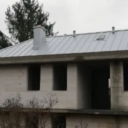 Nowy, srebrny dach z blachy na surowym budynku z pustaków; widoczny komin, okno dachowe i wentylacja.