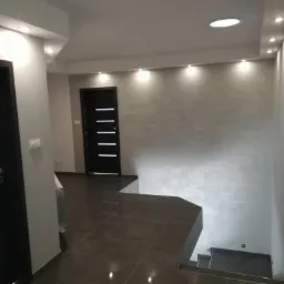 Nowoczesne wnętrze: ciemna podłoga z płytek, szara ściana z subtelną fakturą oświetlona punktowymi lampami, czarne drzwi z poziomymi wgłębieniami, widok na schody.