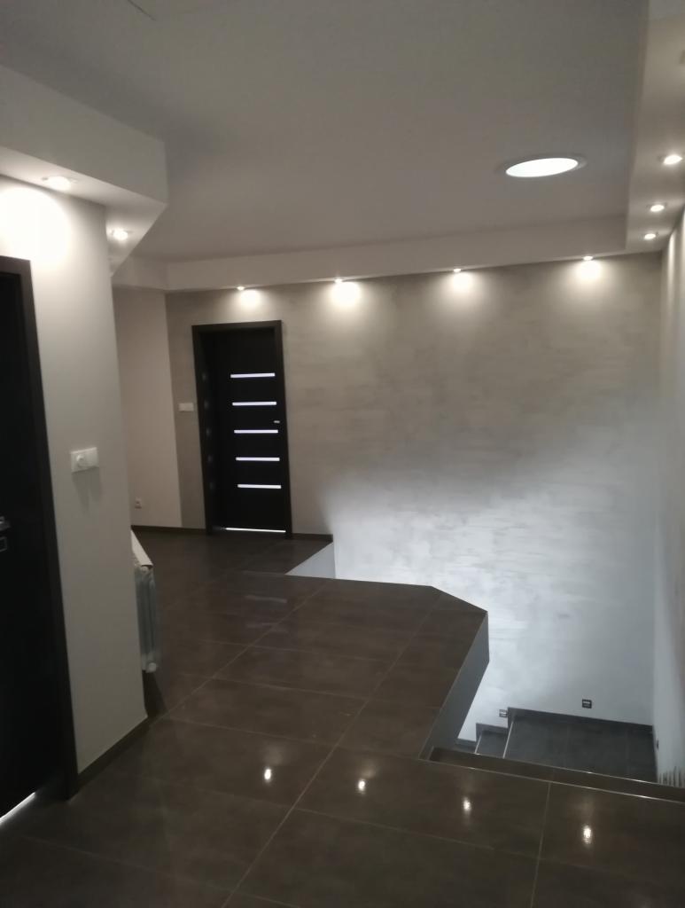 Nowoczesne wnętrze: ciemna podłoga z płytek, szara ściana z subtelną fakturą oświetlona punktowymi lampami, czarne drzwi z poziomymi wgłębieniami, widok na schody.