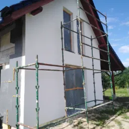 Dom w Lipuszu w trakcie ocieplania, widoczne rusztowanie, fragment elewacji z nałożonym tynkiem oraz niezakończone ocieplenie styropianem.