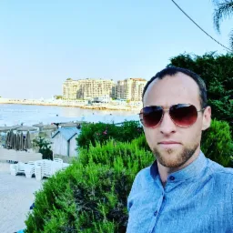 Mężczyzna w okularach na tle plaży z budynkami, morzem i zielenią. Ujęcie z bliska, z wyraźnym pierwszym planem roślinności.