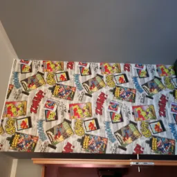 Ściana pokoju dziecięcego wykończona tapetą w komiksowe motywy z postaciami Marvela, widok z dołu, obok fragment łóżka.