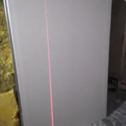 Montaż płyty karton gips: pionowa linia lasera wyznacza idealny pion na szarej płycie, widoczna wełna mineralna w tle.