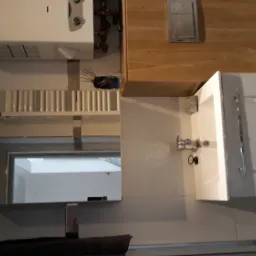 Nowoczesna łazienka z białą szafką pod umywalką, lustrem z oświetleniem, bojlerem i drewnianą zabudową, widoczne fragmenty instalacji hydraulicznej.