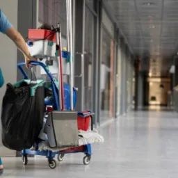 Pracownik w niebieskim uniformie pcha wózek z akcesoriami do sprzątania wzdłuż korytarza biurowego z przeszklonymi ścianami.