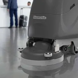 Szara, automatyczna maszyna do mycia podłóg marki Numatic na tle polerowanej podłogi biurowej i recepcji z dwoma osobami w tle.