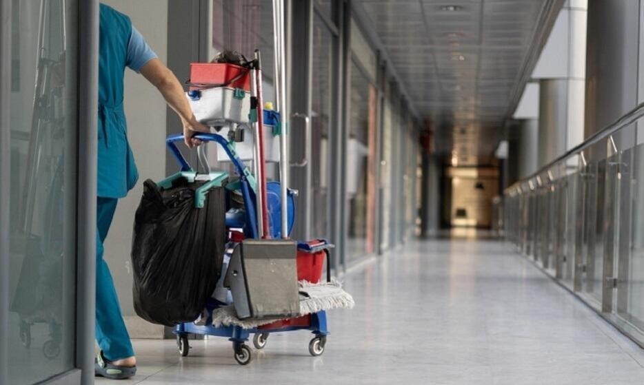 Pracownik w niebieskim uniformie pcha wózek z akcesoriami do sprzątania wzdłuż korytarza biurowego z przeszklonymi ścianami.