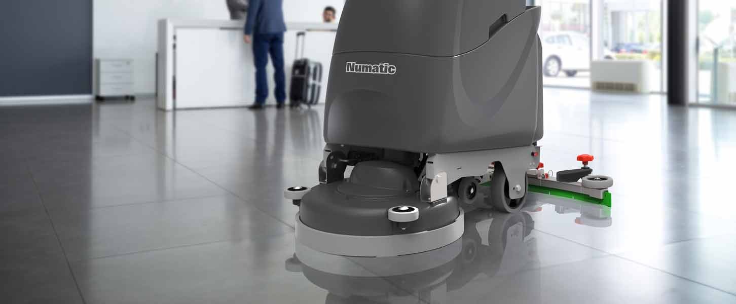 Szara, automatyczna maszyna do mycia podłóg marki Numatic na tle polerowanej podłogi biurowej i recepcji z dwoma osobami w tle.