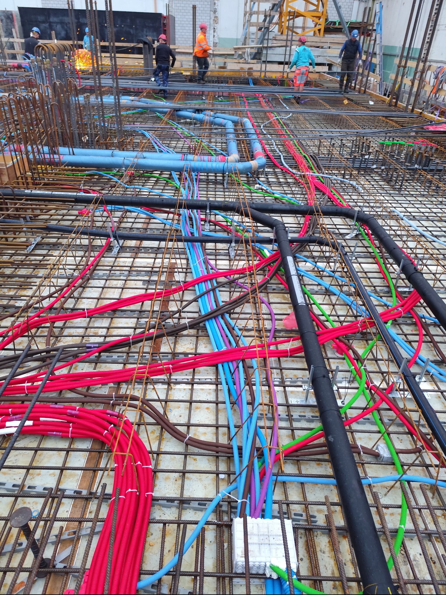 Zbrojenie płyty fundamentowej z instalacją: rury w kolorach czerwonym, niebieskim, zielonym i czarnym, ułożone na siatce zbrojeniowej. W tle pracownicy budowlani.