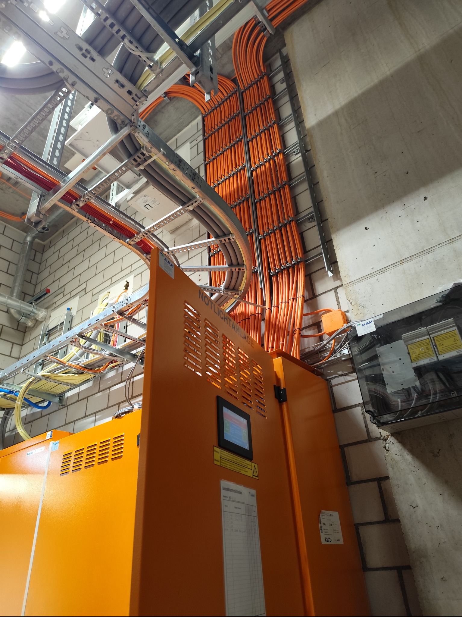 Pomarańczowe kable ułożone wzdłuż ściany, prowadzone na metalowych konstrukcjach. Widoczna szafa sterownicza i instalacje w budynku z ceglanymi ścianami.