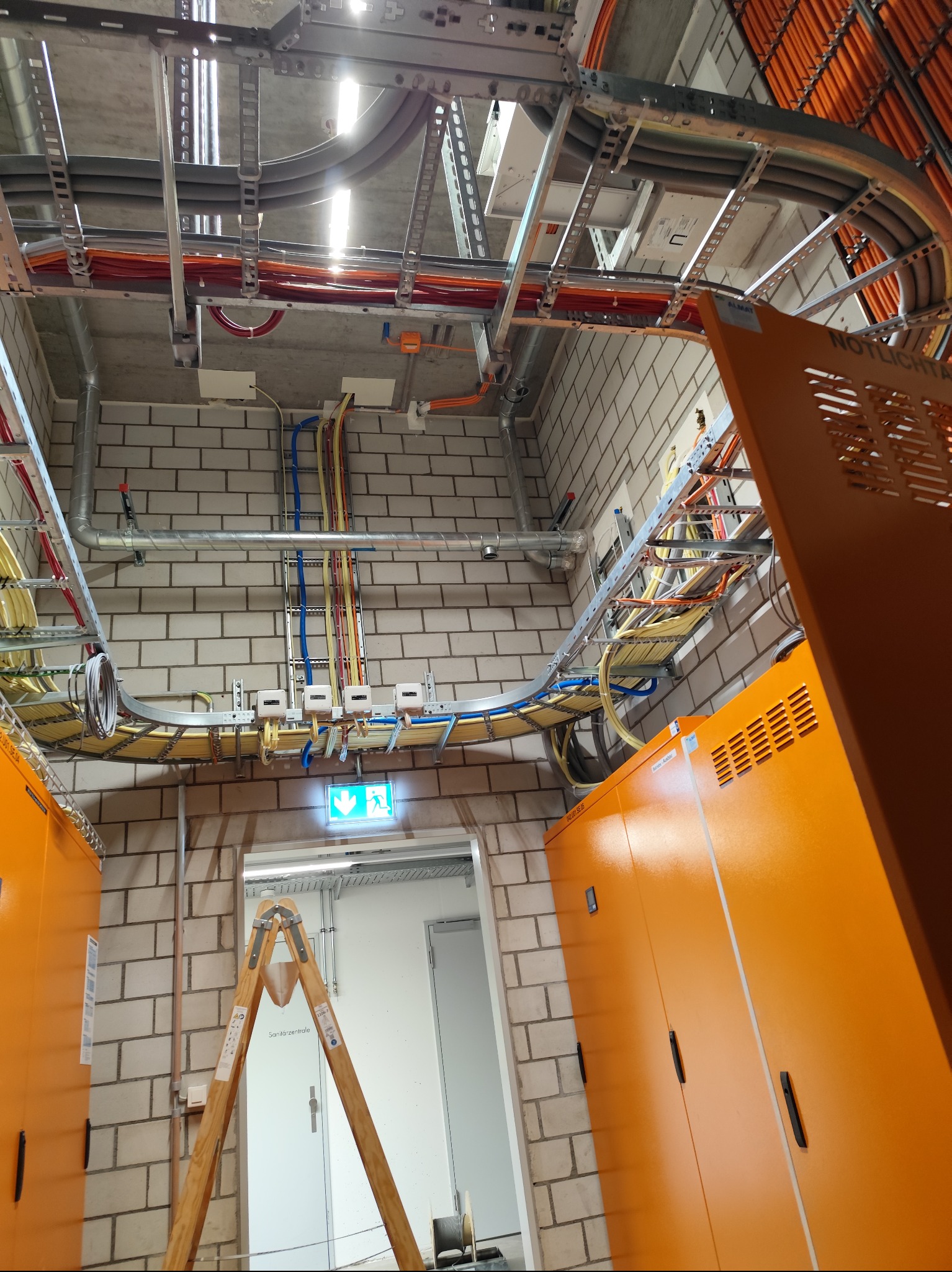 Rozbudowana instalacja kablowa z pomarańczowymi szafami, widoczna z dołu. Kable ułożone na metalowych korytach pod sufitem w pomieszczeniu z ceglanymi ścianami.