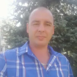 Mężczyzna w niebieskiej koszuli w kratę pozuje na tle drzewa iglastego i budynku z oknami. Widoczny srebrny łańcuszek na szyi.