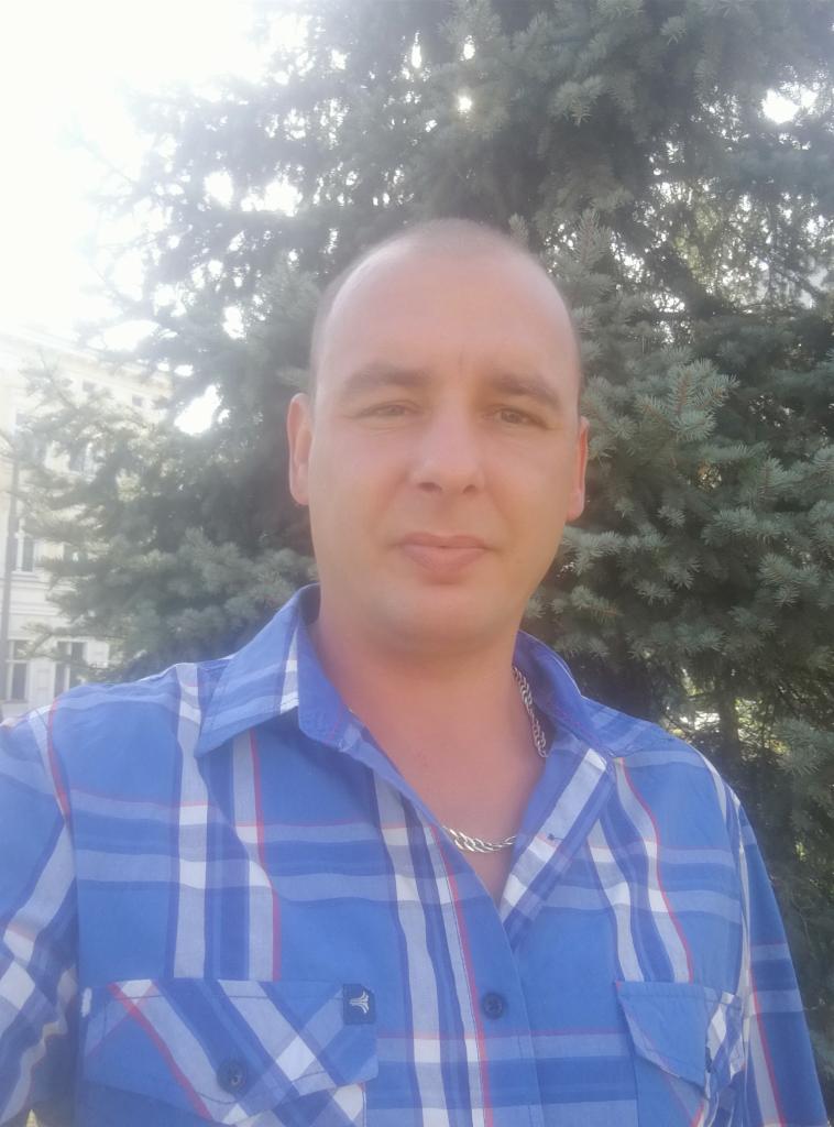 Mężczyzna w niebieskiej koszuli w kratę pozuje na tle drzewa iglastego i budynku z oknami. Widoczny srebrny łańcuszek na szyi.