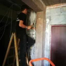 Elektryk na drabinie, mocuje kable elektryczne w rurach na surowym betonowym suficie w trakcie remontu, w tle widoczny otwór drzwiowy.