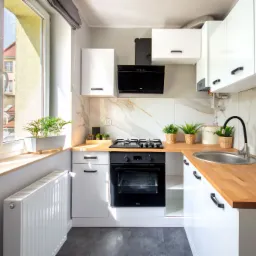 Jasna, nowocześnie urządzona kuchnia z białymi szafkami, drewnianym blatem, czarną zabudową piekarnika i płyty grzewczej, oraz marmurowymi płytkami na ścianie, doświetlona naturalnym światłem...