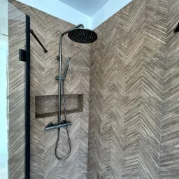 Wykończona kabina prysznicowa z deszczownicą w kolorze grafitowym i ścianami wyłożonymi płytkami imitującymi drewno ułożonymi we wzór jodełki. W tle widoczny fragment niedokończonej ściany.