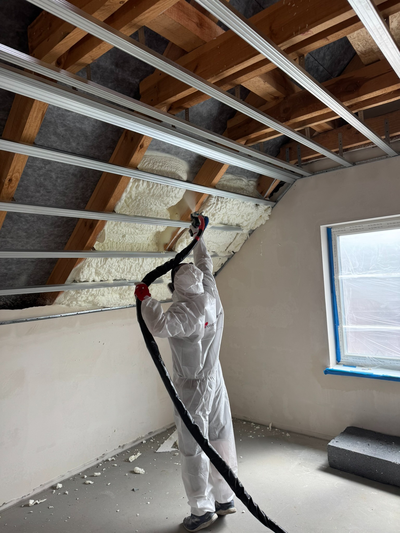 Aplikacja pianki PUR na poddaszu przez pracownika w kombinezonie ochronnym. Widoczne drewniane elementy konstrukcji dachu i metalowe profile. Okno zabezpieczone folią.