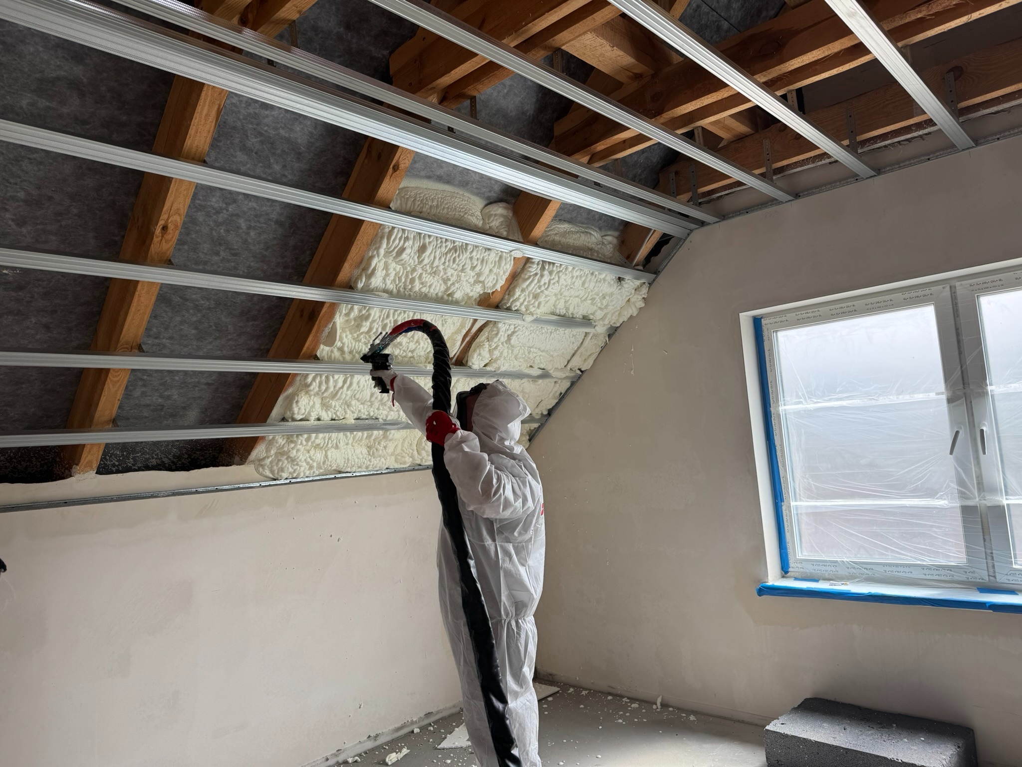 Aplikacja pianki PUR na poddaszu przez pracownika w kombinezonie ochronnym. Widoczne drewniane elementy konstrukcji dachu i metalowe profile. Okno zabezpieczone folią.