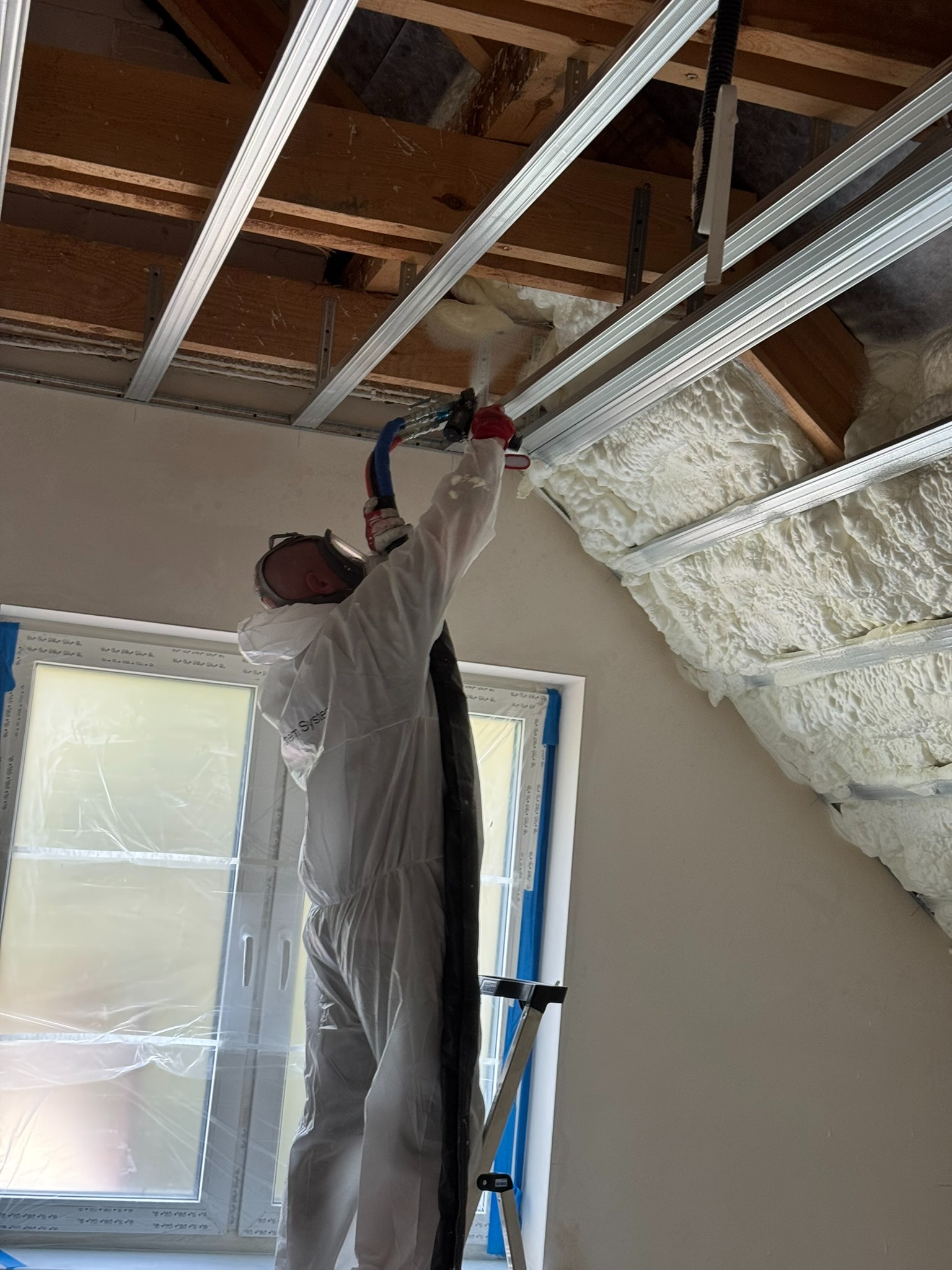 Pracownik w kombinezonie aplikuje piankę PUR na poddaszu, widoczne drewniane elementy konstrukcji dachu i okno zabezpieczone folią. Izolacja natryskowa.